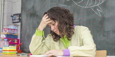 stressed teacher with head in hand sitting at desk in classroom