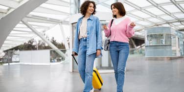 two smiling women walking through airport together with luggage