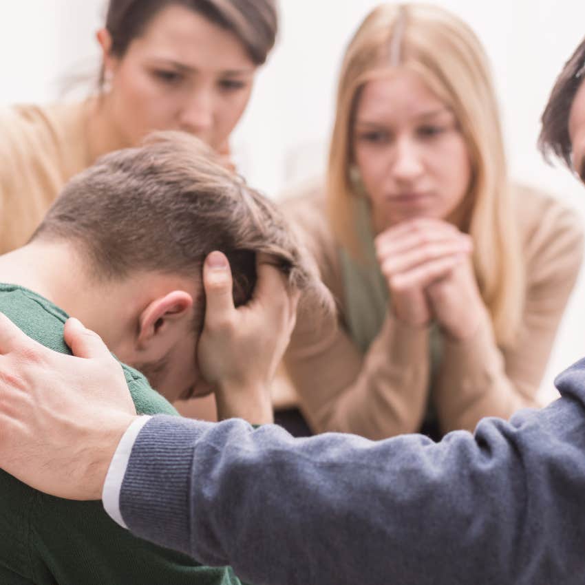 Close-up of a devastated young man holding his head in his hands and friends supporting him during group therapy