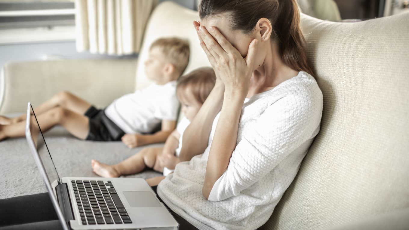sad, stressed mom on laptop sitting on couch next to kids