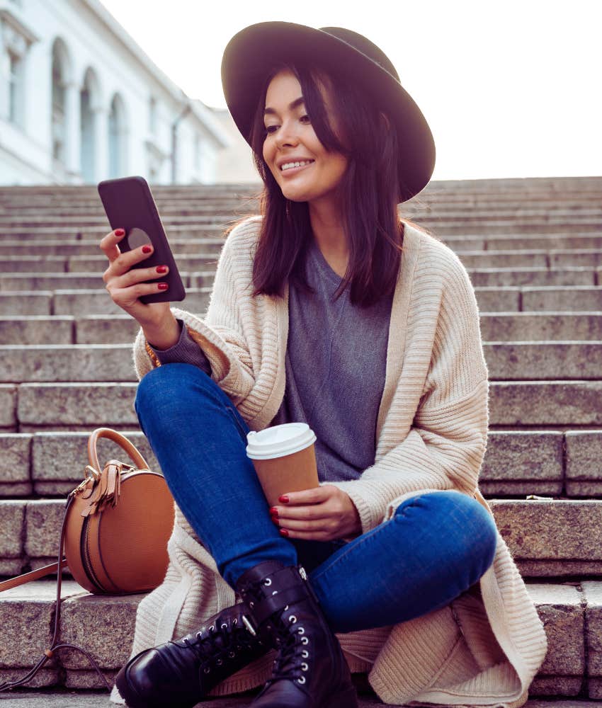 millennial woman holding a coffee sitting on steps texting