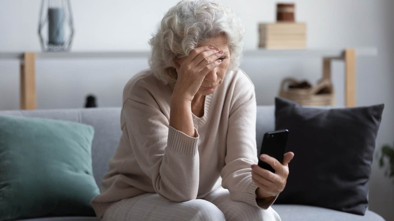 upset middle aged woman looking at phone screen sitting on couch at home