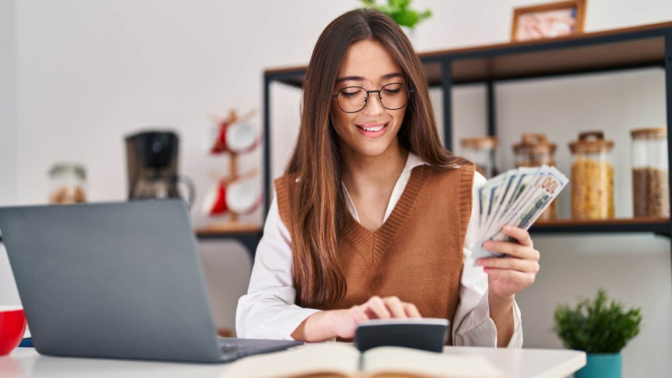 woman counts her saved up cash at home in front of laptop