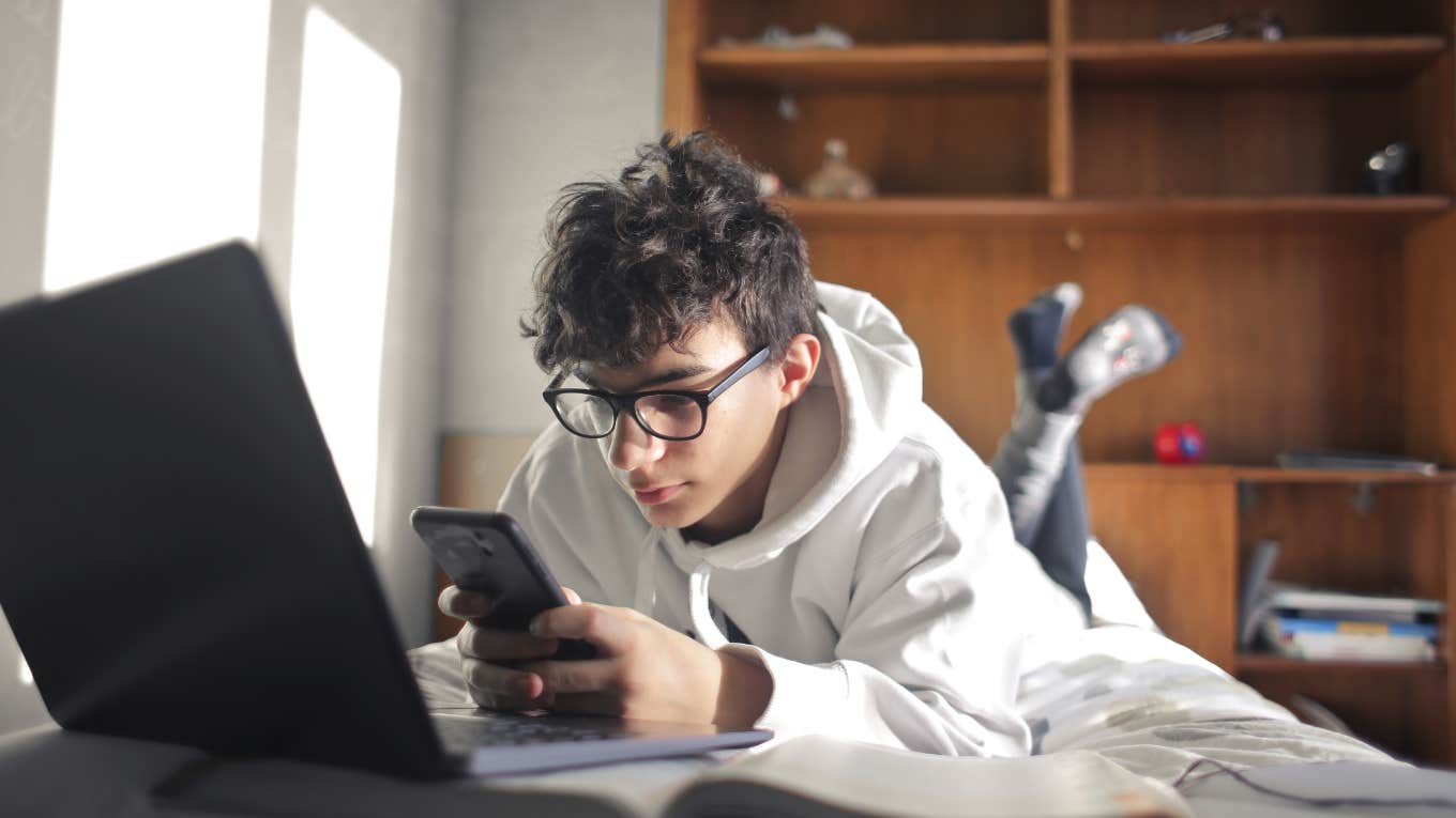 young boy studying while lying on the bed using computer and smartphone
