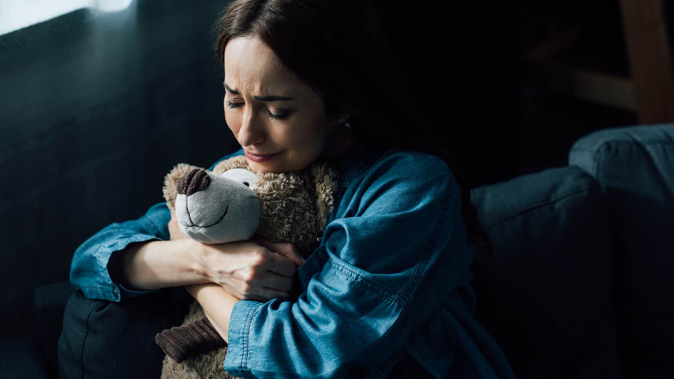 sad brunette woman with closed eyes holding teddy bear in living room