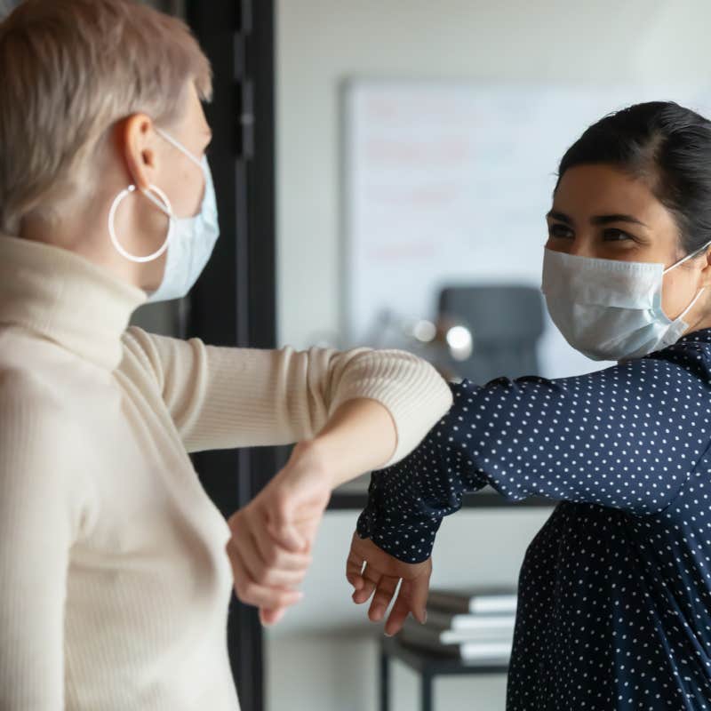 female colleagues wearing protective face masks greeting bumping elbows at workplace