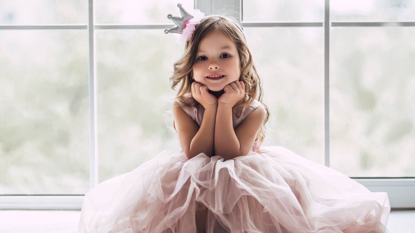 Little cute girl in beautiful dress is sitting near the window at home.