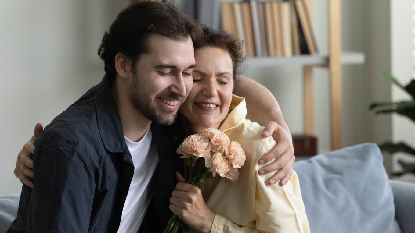 mom embracing grown son man while holding flowers.