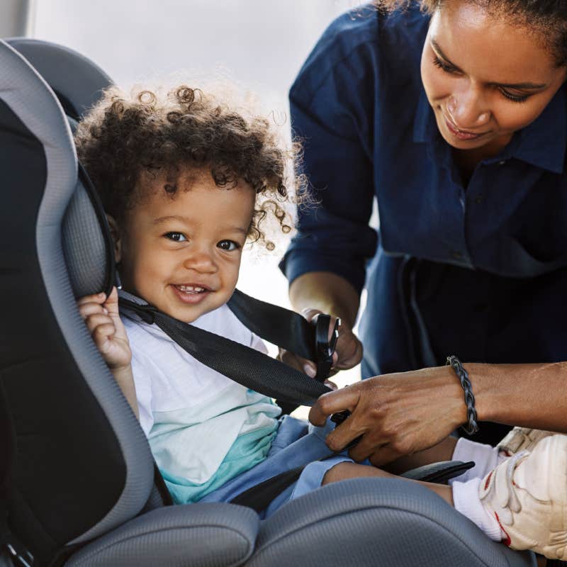 Side view of a happy little boy looking at camera while his mother buckling him in a car seat