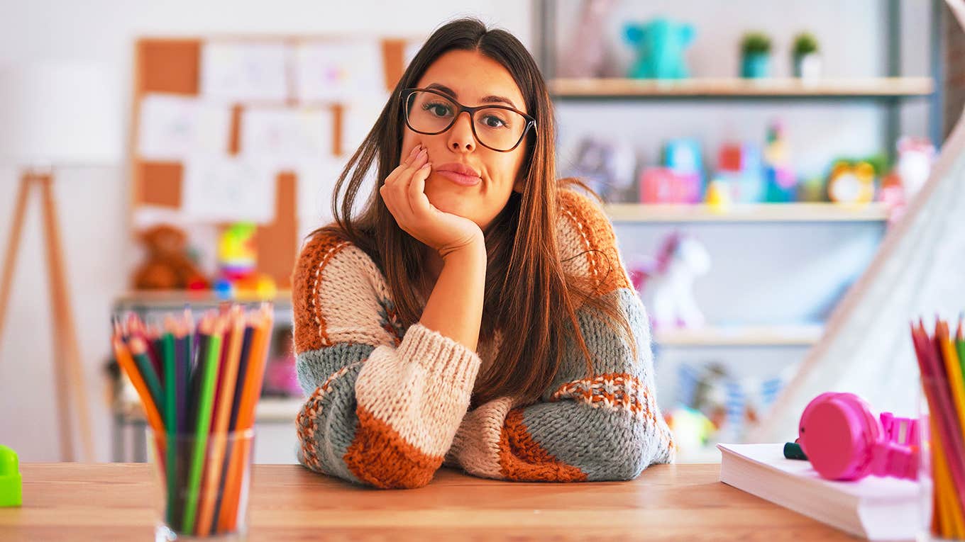 teacher woman wearing sweater and glasses sits at desk looking frustrated and annoyed with crossed arms.