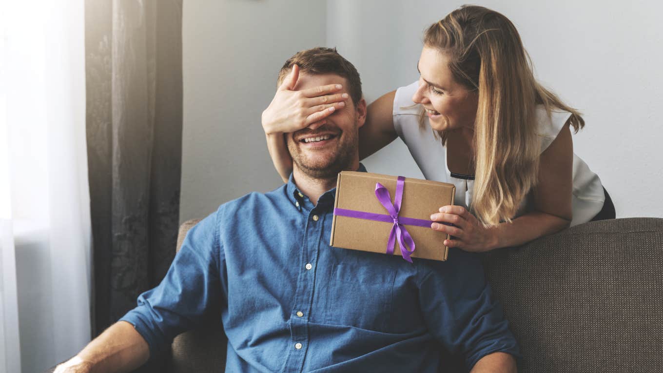 wife covering husband's eyes while handing him wrapped present