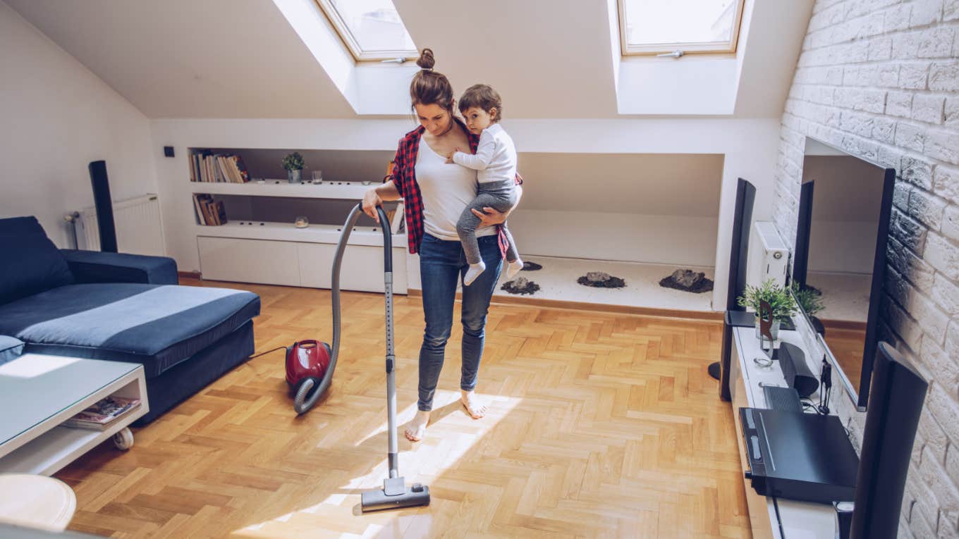 woman cleans and vacuums living room while holding boy in her arms