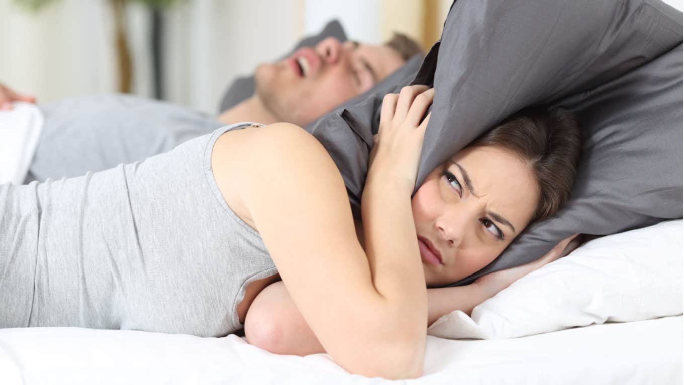 uncomfortable woman holding pillow over ears in bed next to man snoring 