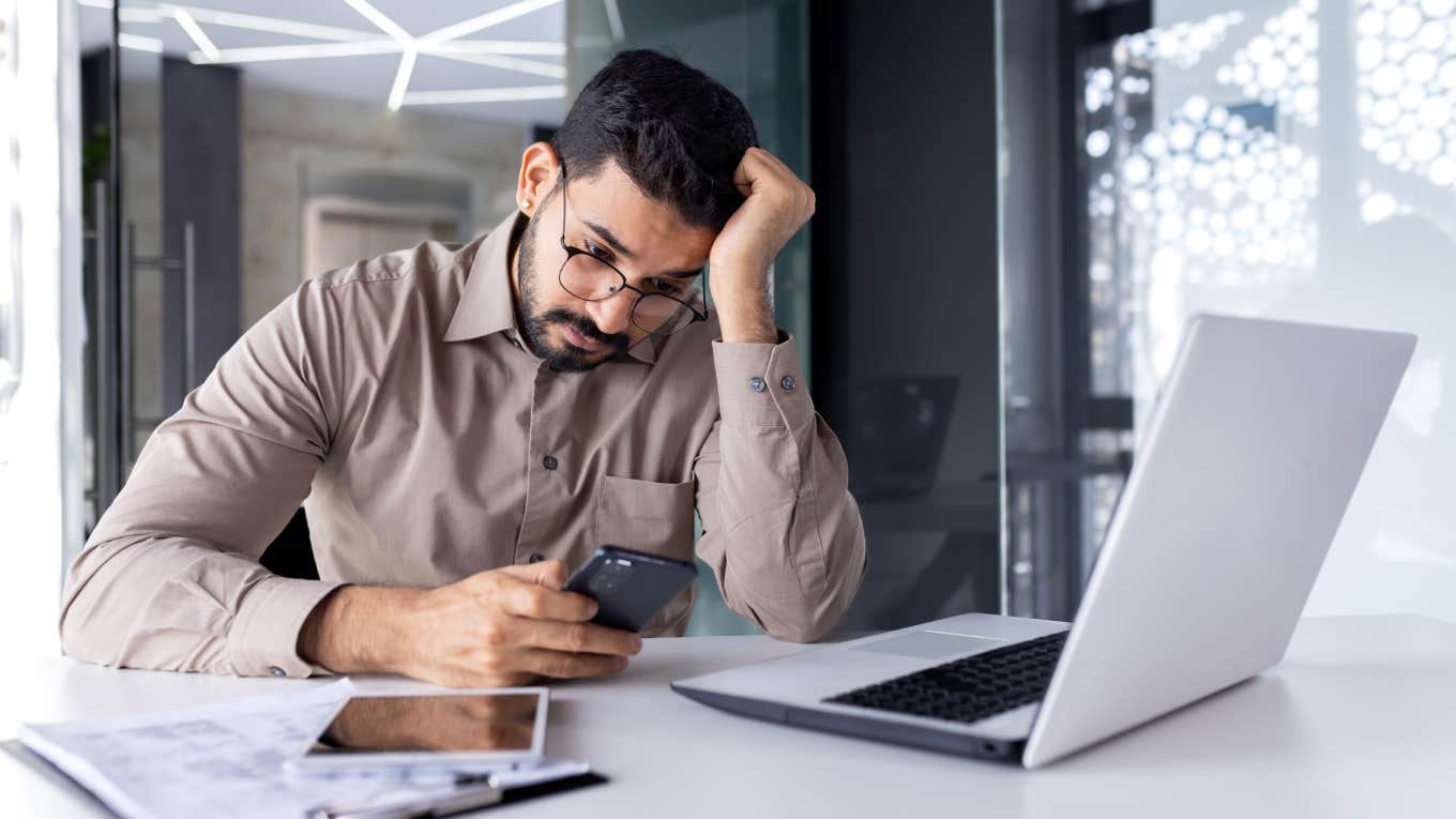 upset businessman at workplace inside office reading bad news on phone