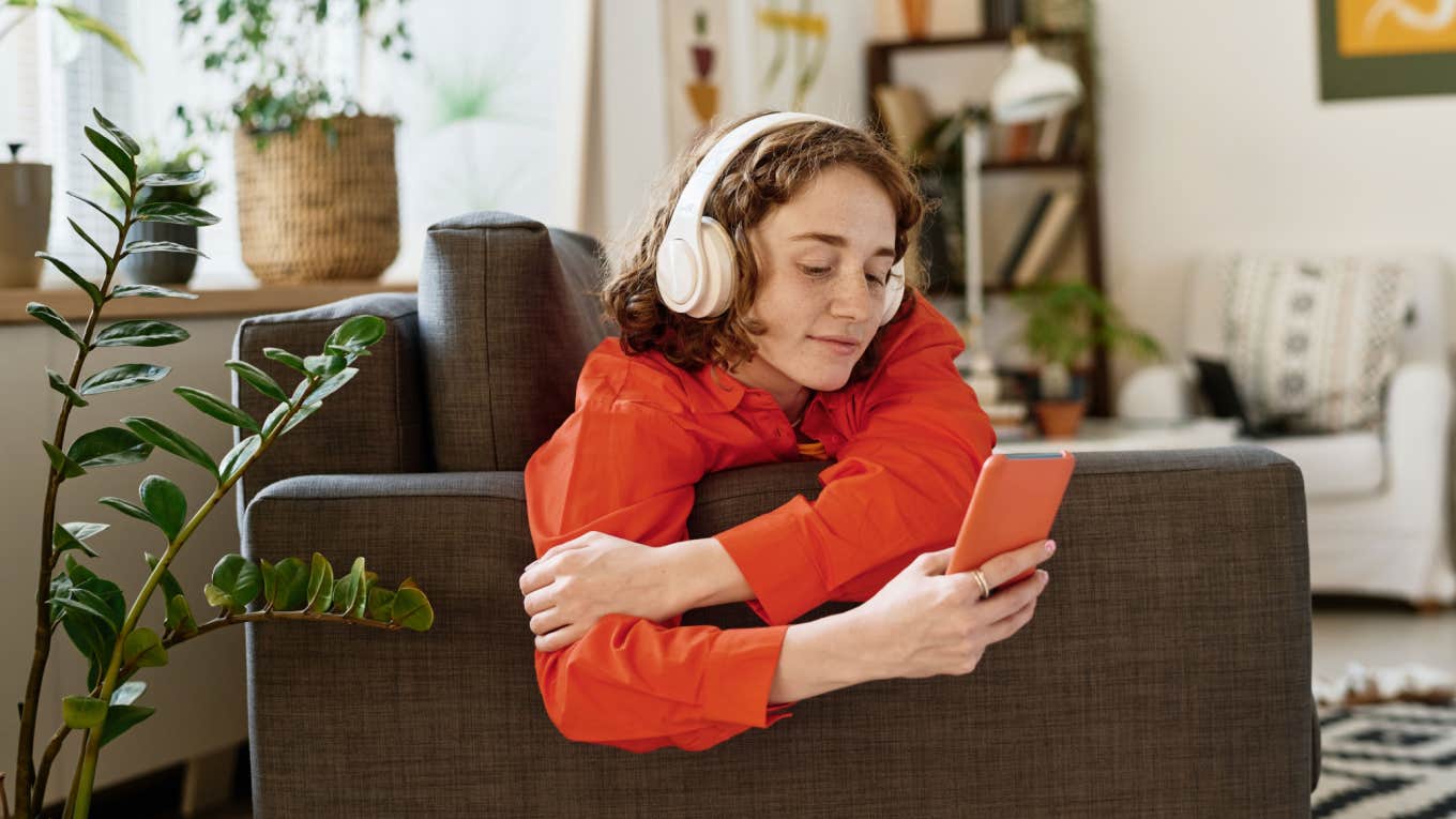 young woman with headphones on sits on couch in living room while on phone