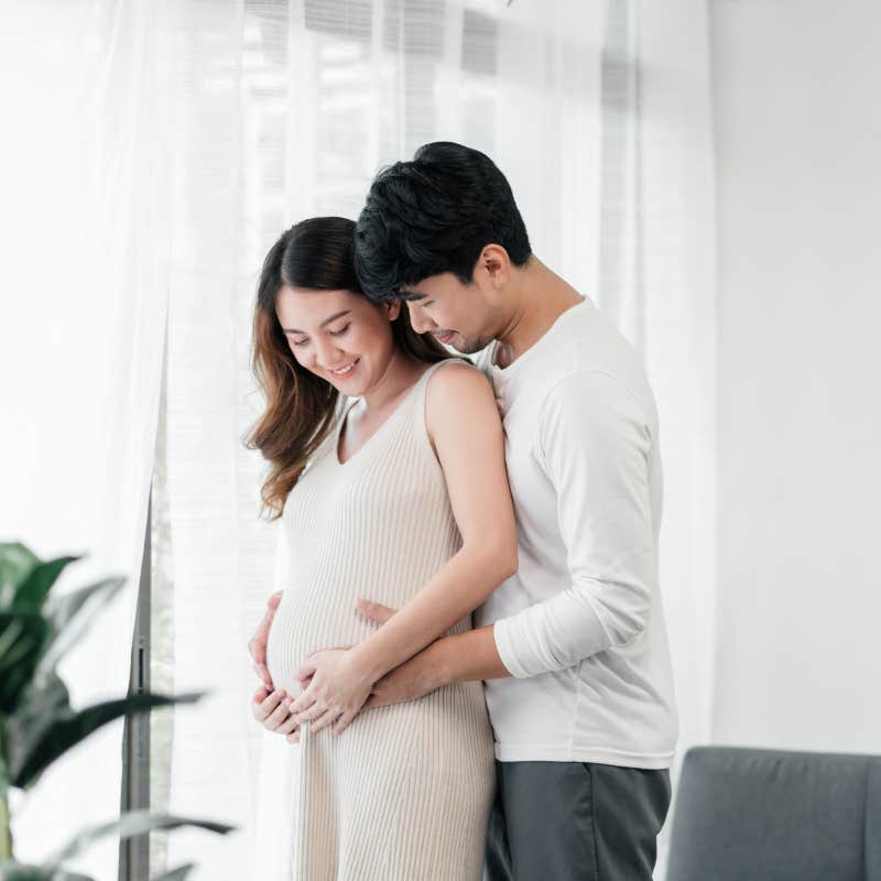 husband embracing his pregnant wife next to the window in the living room.