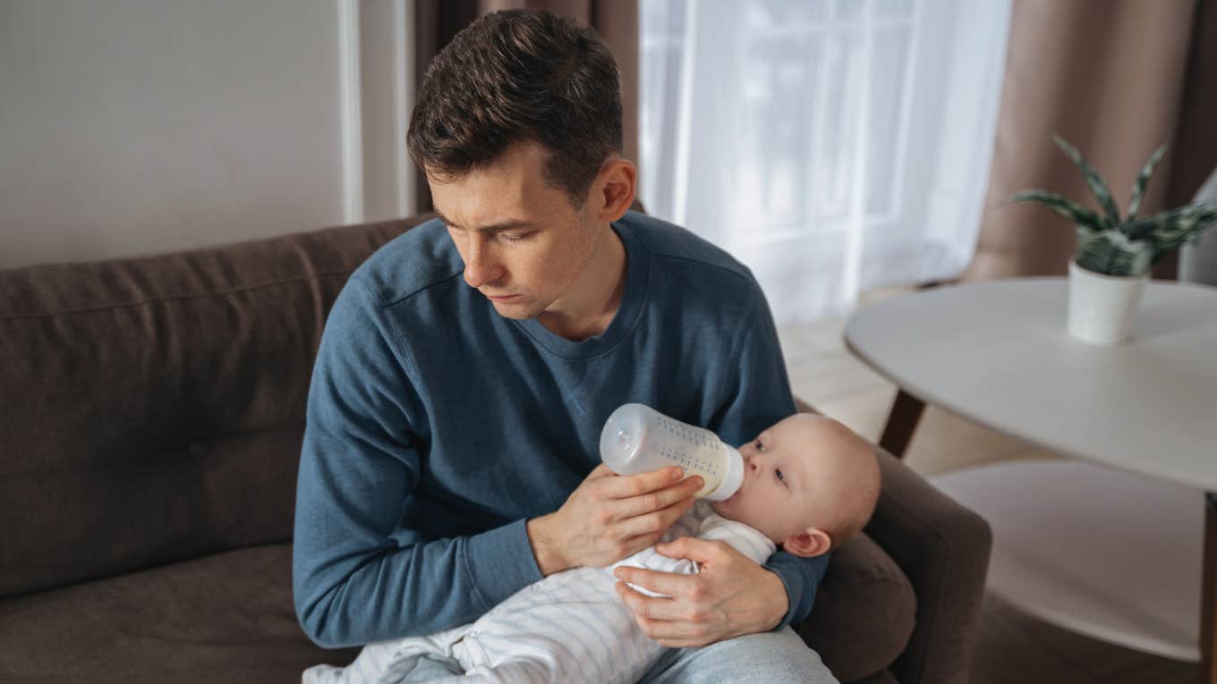 Unhappy young father feeding newborn baby with milk bottle on couch at home