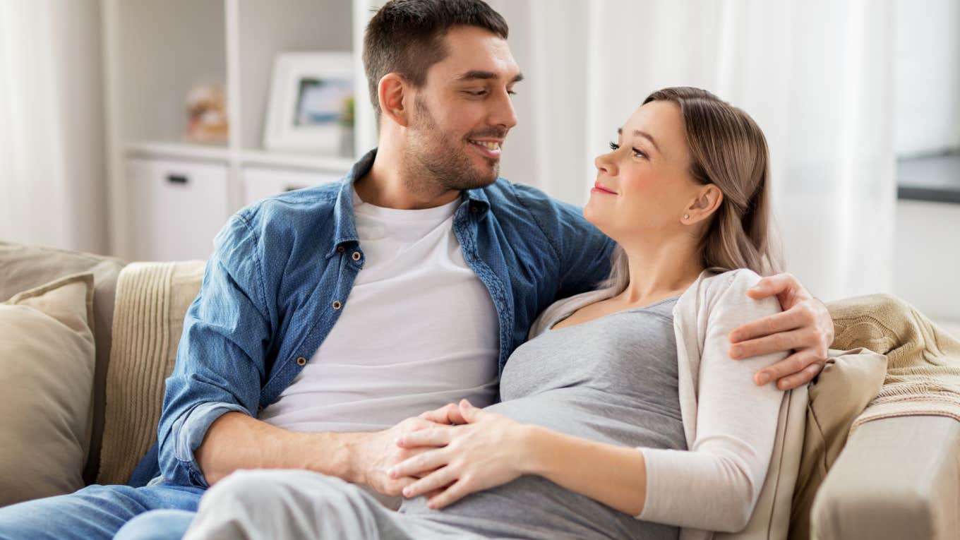 husband hugging pregnant wife and rubbing belly while sitting on couch