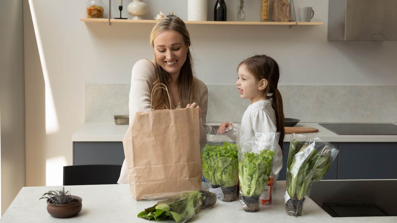 young mother and joyful cute small kid daughter unpacking fresh salad leaves and organic greenery from carton package