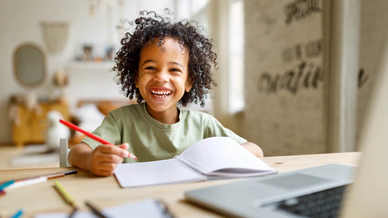 smiling little boy sitting in classroom with book open and pencil in hand