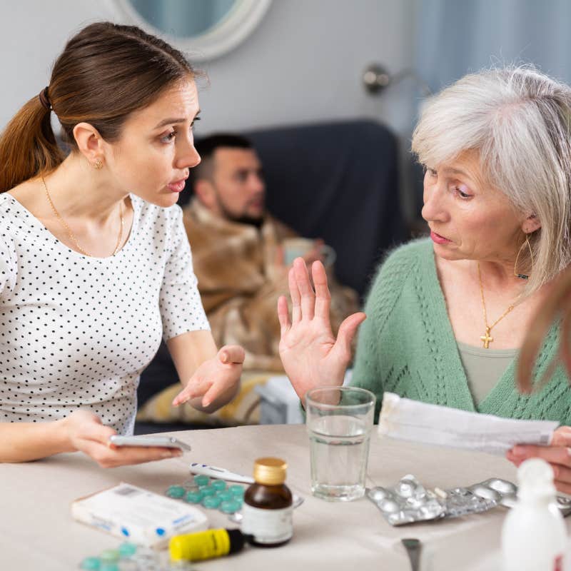 woman arguing with mother-in-law sitting at table with husband on couch behind them