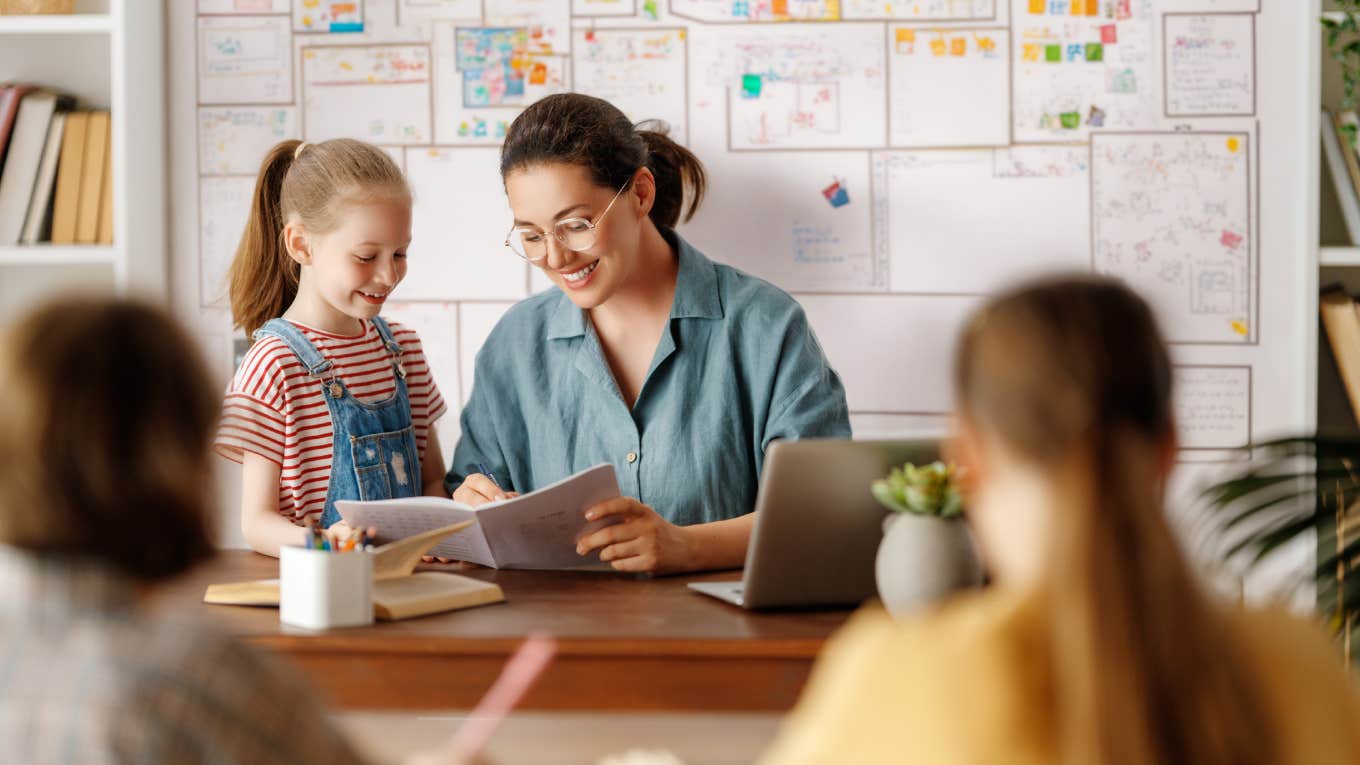 child talking to teacher sitting behind desk in classroom