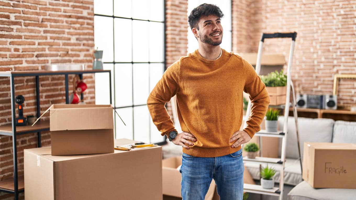 Young man smiling and standing in new home surrounded by cardboard boxes