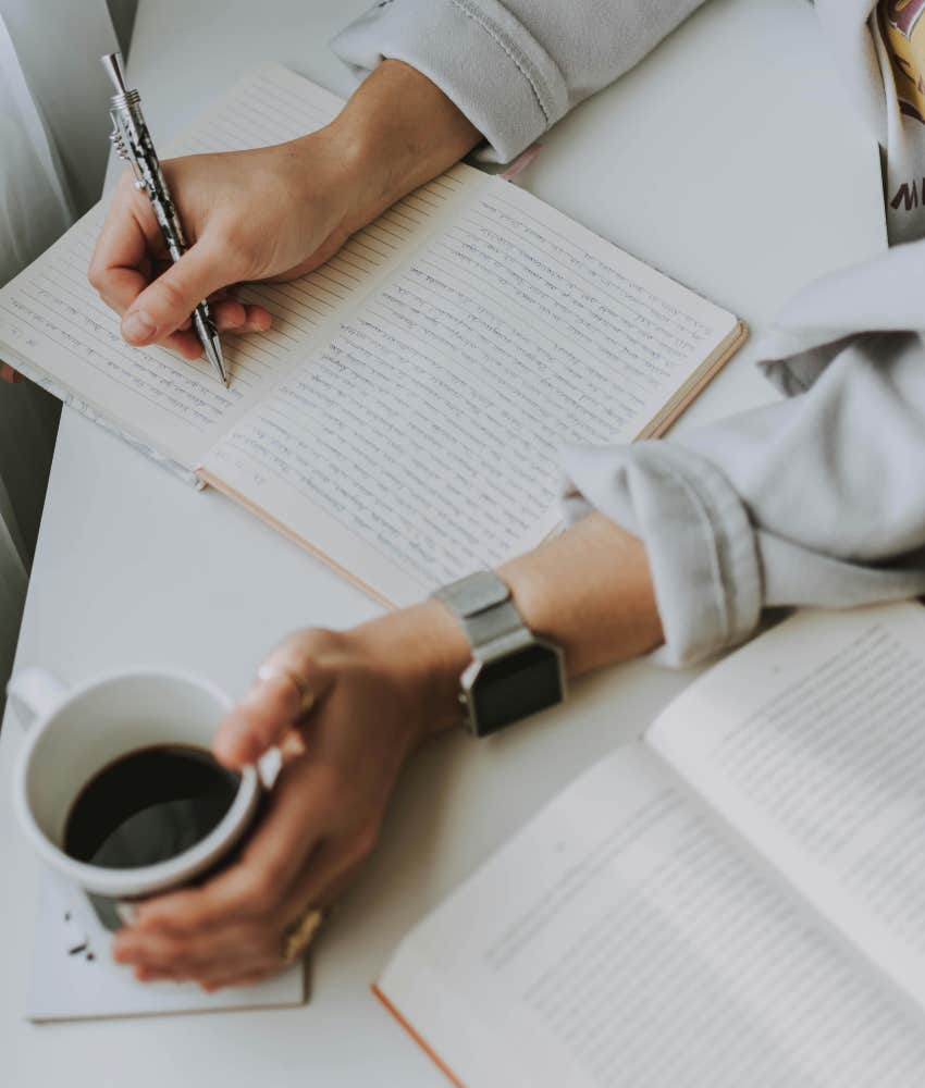 person writing in journal holding coffee mug