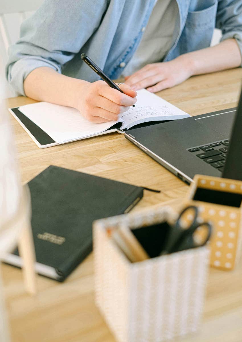 person writing in a notebook in front of a laptop