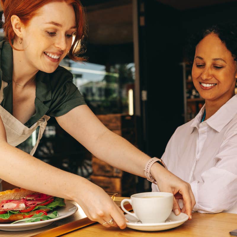 Happy young waitress serving a customer a sandwich and a cup of coffee in a modern cafe