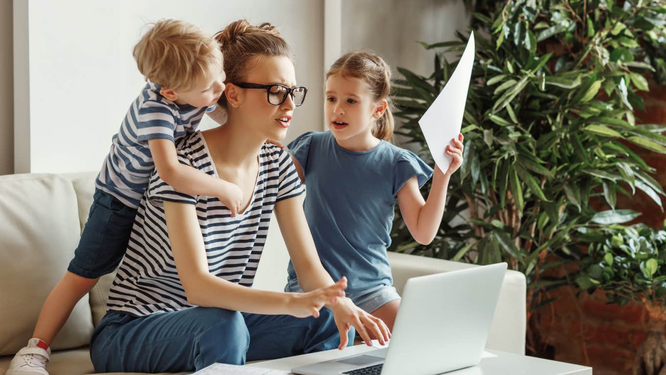 stressed and exhausted mom working on laptop with kids around her