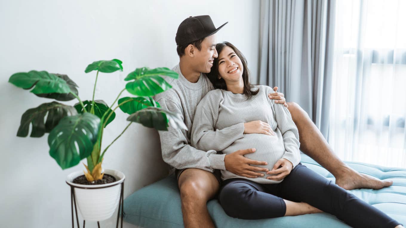 man and pregnant woman embrace each other while sitting by window at home