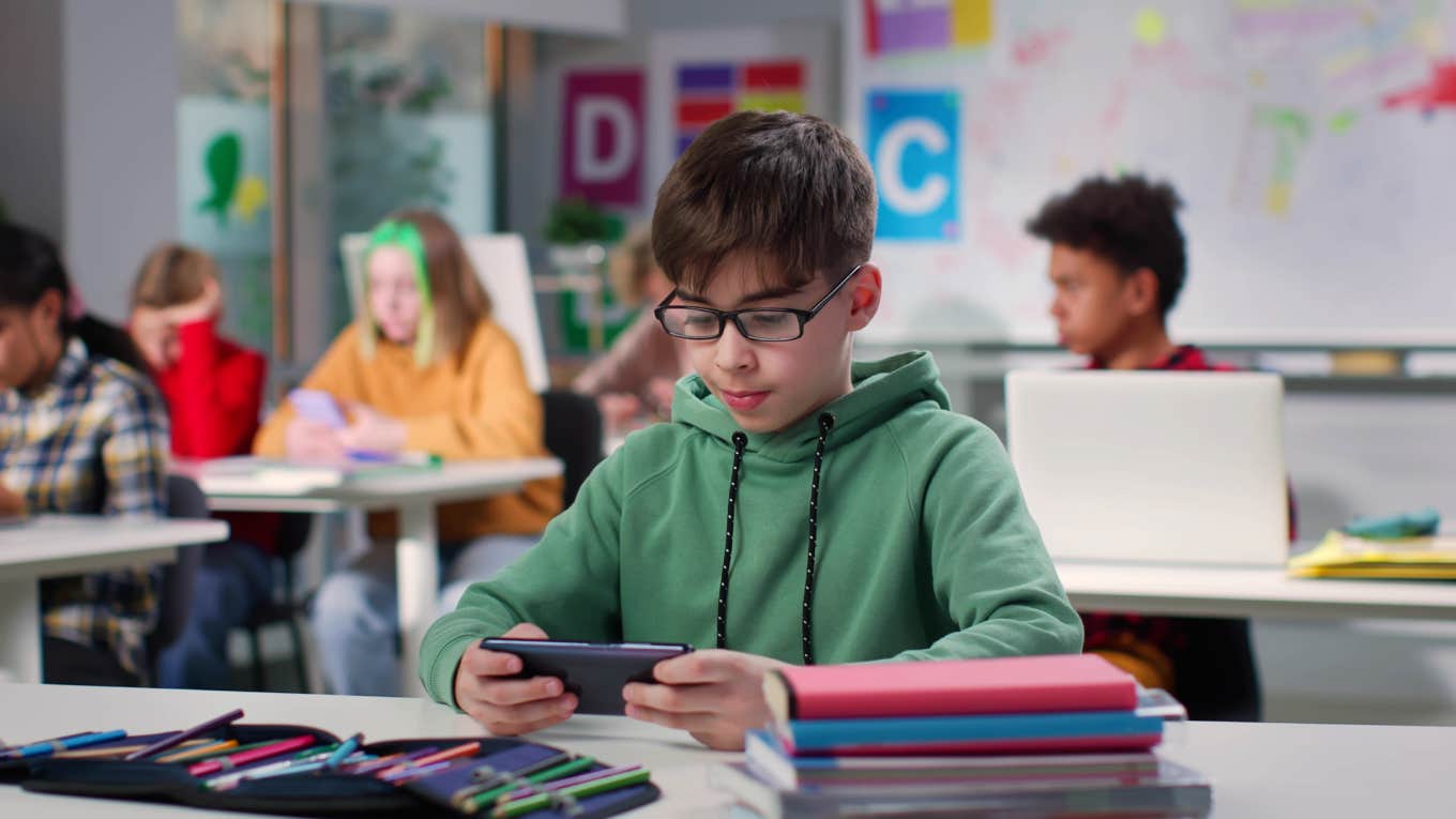 teen boy watching videos on smartphone sitting at desk in class