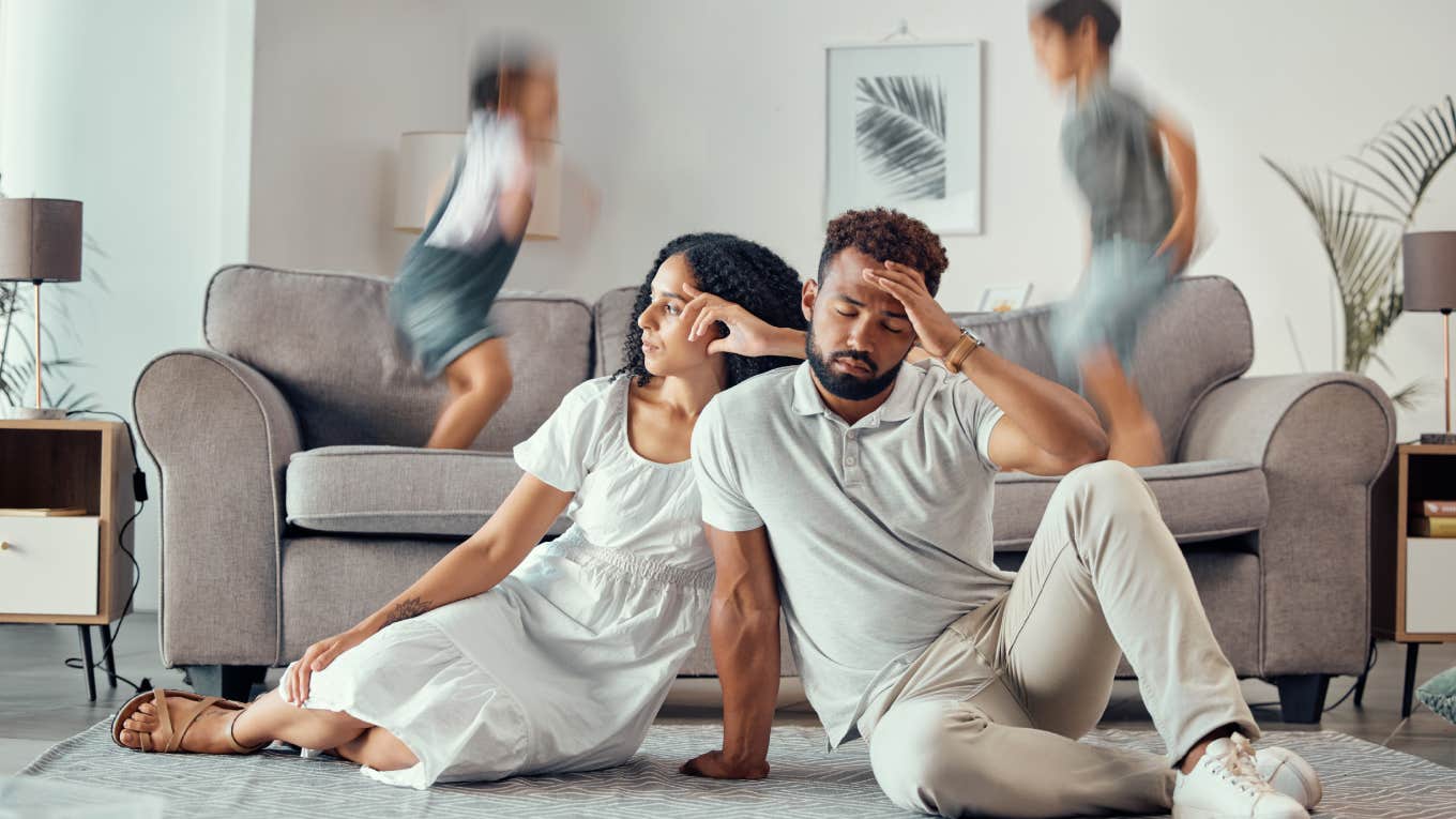 stressed out parents sitting on the floor while kids jump on couch behind them