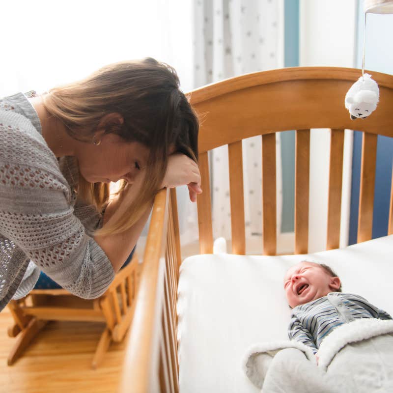 tired mother leaning over side of crib while newborn baby cries