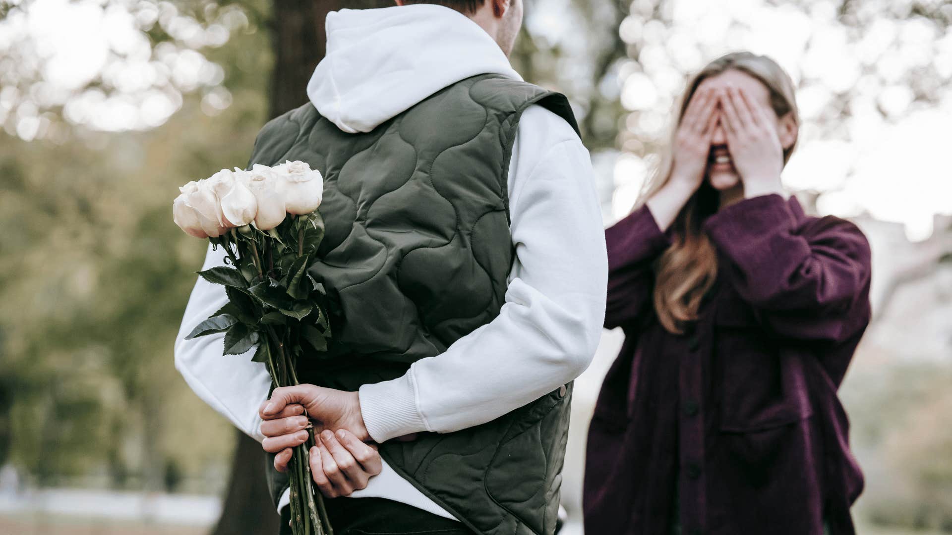 man hiding flowers behind back while woman covers her eyes