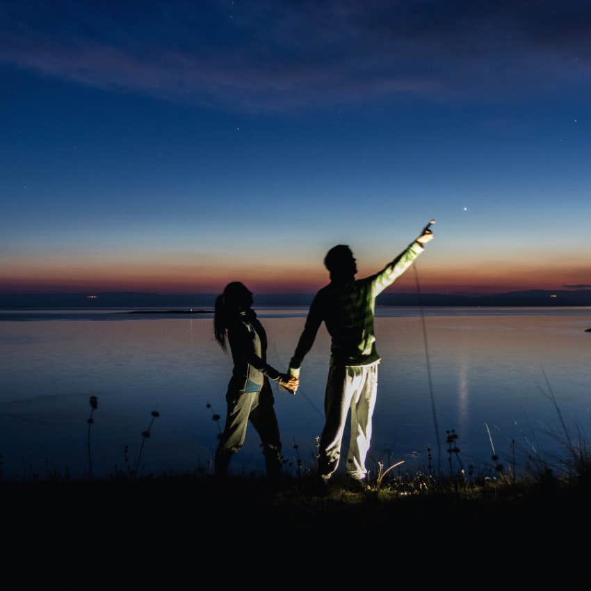 man holding woman's hand pointing up to star shining in night sky at dusk on the beach