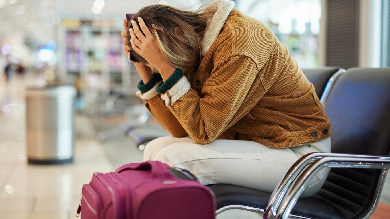 upset woman sitting with head in hands at airport with luggage