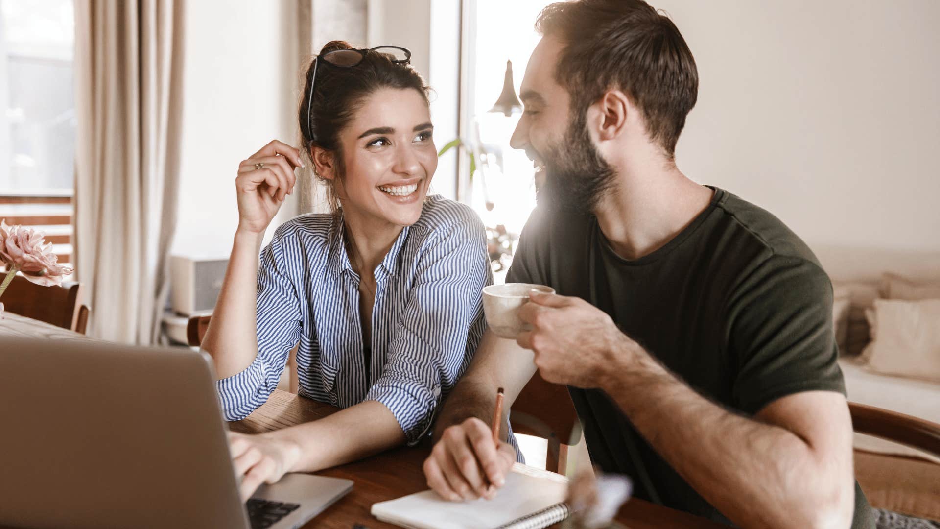 couple chatting and smiling while man drinks hot drink and writes something down