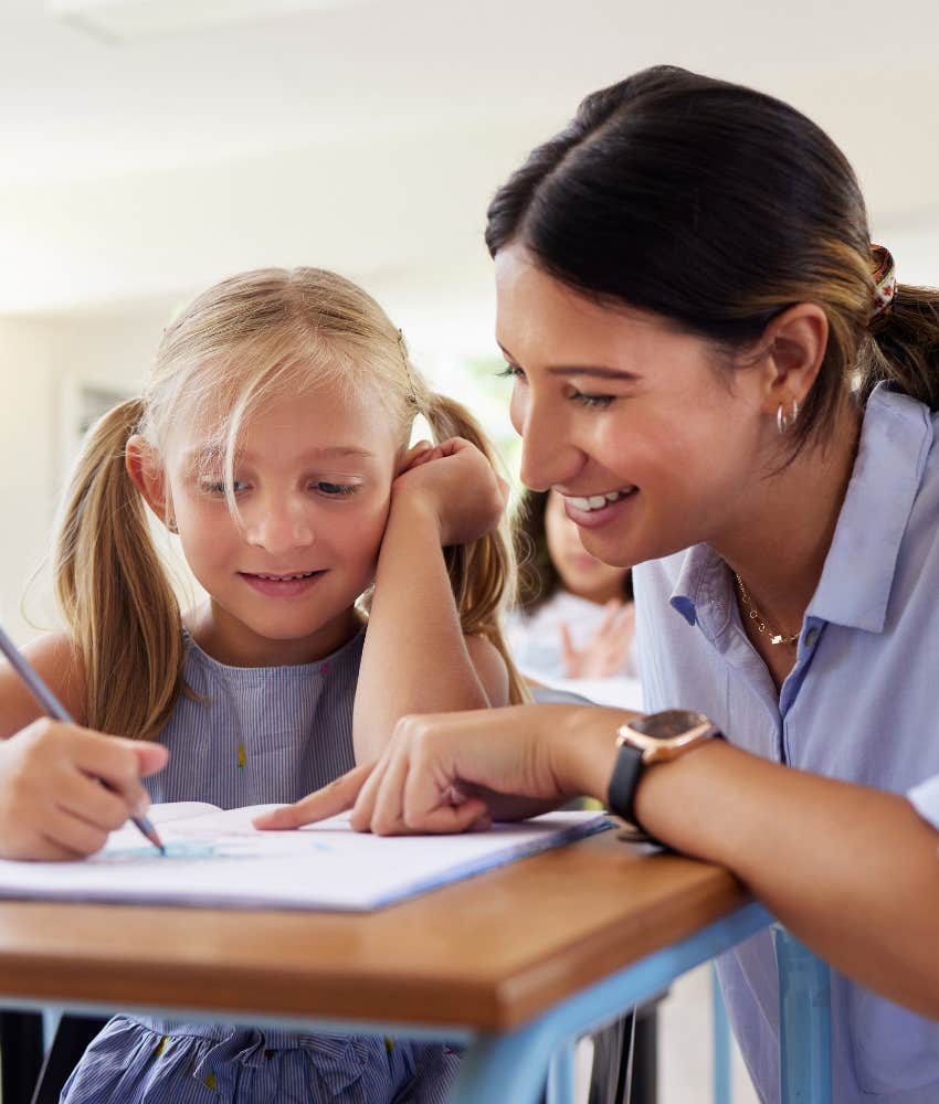 smiling teacher helping little girl with pigtails complete assignment