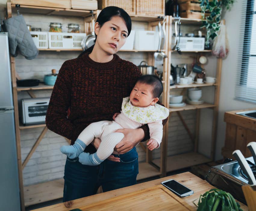 tired and overwhelmed stay-at-home mom holding infant baby in kitchen while staring into the distance 