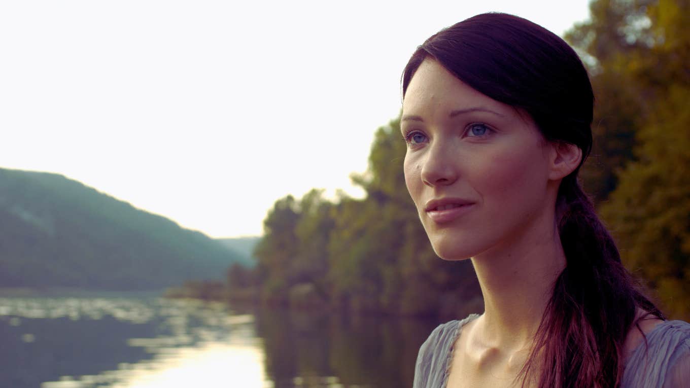 brunette standing by a river with a slight grin