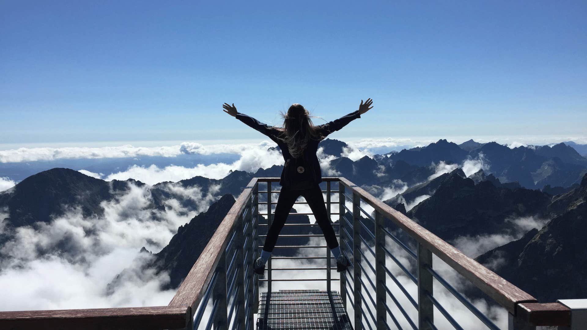 woman with raised arms on bridge over mountains