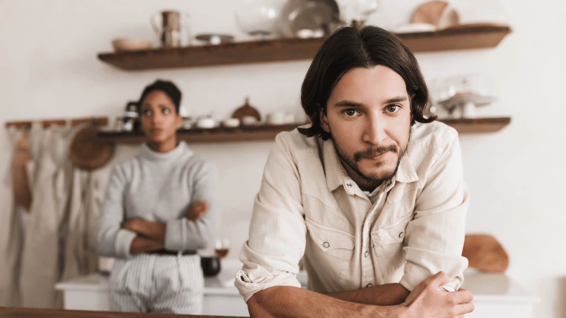 Disgruntled man leans on table while woman is dissatisfied behind him leaning against wall