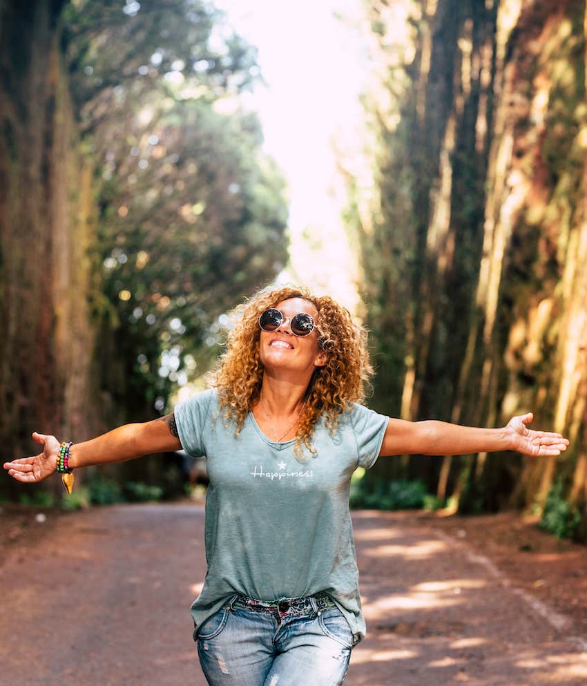 Woman walks down narrow path in nature with arms outstretched to take it all in