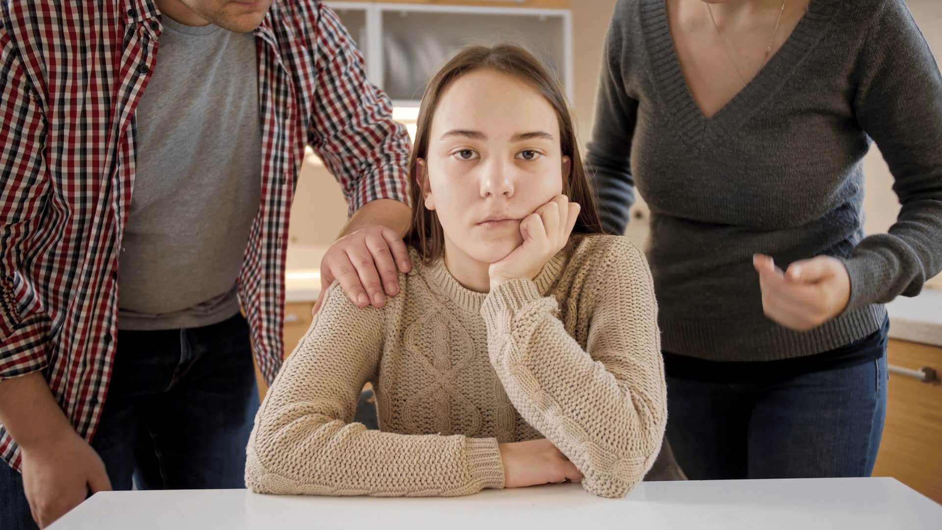 Teenager looks annoyed with each parent placing a hand on her shoulders