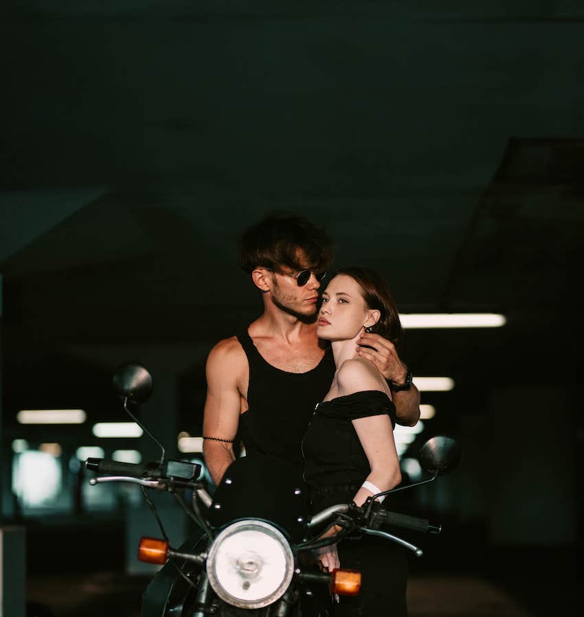 Man and woman side hug while he wears sunglasses and is on a motorcycle 