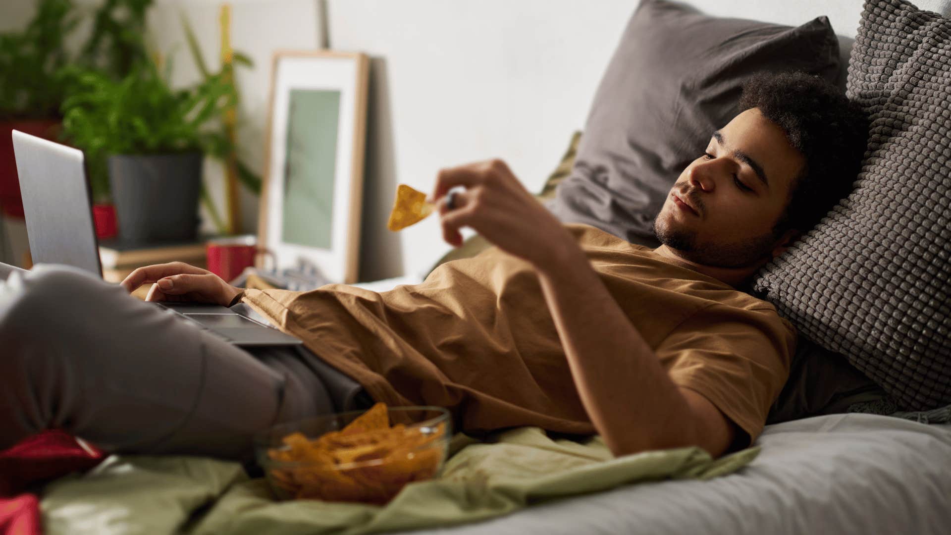 man lying on bed looking at laptop while eating chips