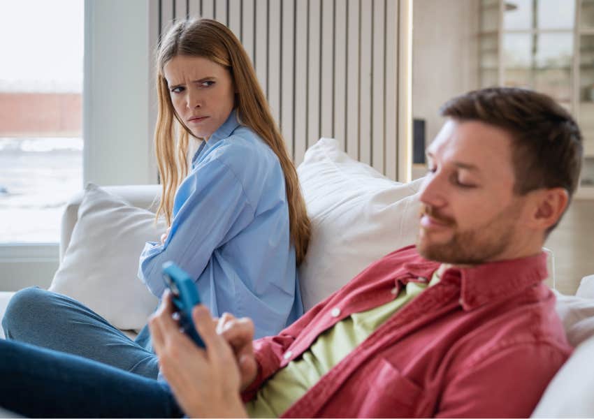 concerning wife glancing at husband smiling at phone on couch at home