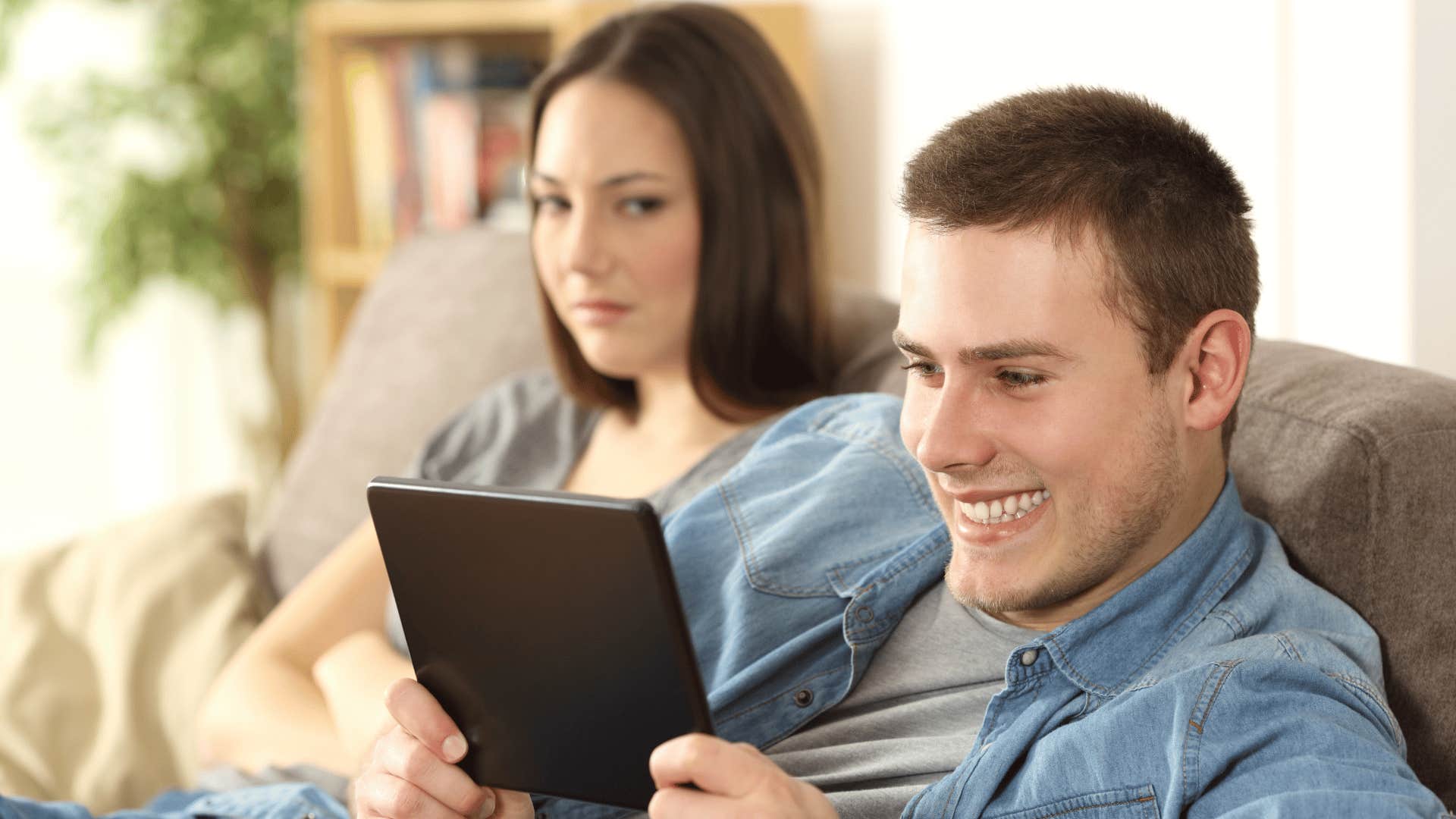 man on tablet ignoring woman while she stares at him annoyed